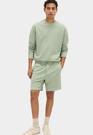 Hombre vistiendo sudadera de manga larga verde claro y pantalones cortos a juego, con calcetines beige y zapatillas, de pie con una mano en el bolsillo.