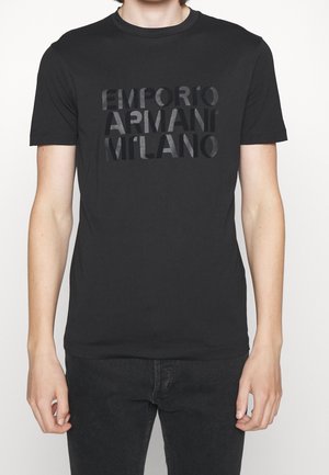 Młoda osoba nosząca czarny t-shirt z krótkim rękawem z wytłoczonym napisem "EMPORIO ARMANI MILANO" na piersi, w połączeniu z czarnymi spodniami.