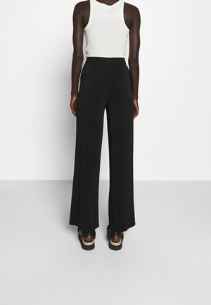 Personne portant un pantalon noir large, un top blanc sans manches et des sandales à plateforme noires, debout face à un mur blanc uni.
