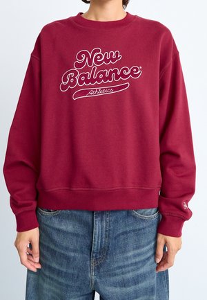 Person iført en mørkerød sweatshirt med teksten "New Balance Athletics" og blå jeans, stående mod en ensartet lys baggrund.