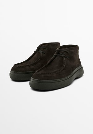 Braune Wildleder-Ankle-Boots mit runder Zehenpartie, Schnürdesign und einer dicken, strukturierten grünen Gummisohle. Nähte entlang der Kante.