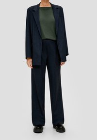 Veste à carreaux bleu marine foncé avec de larges revers, associée à un t-shirt vert ajusté et à un pantalon assorti ample, le tout porté avec des chaussures noires.