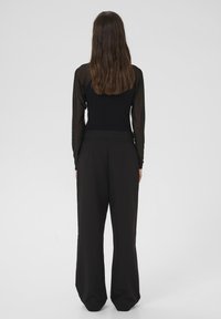 Mujer con cabello largo y castaño de espaldas, vestida con una blusa negra de manga larga y transparente y pantalones negros holgados, sobre un fondo claro y liso.