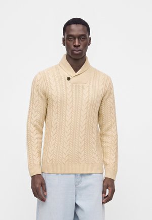 Homme portant un pull beige en maille torsadée avec un col haut et des détails de boutons, associé à un jean bleu clair, debout devant un fond uni.