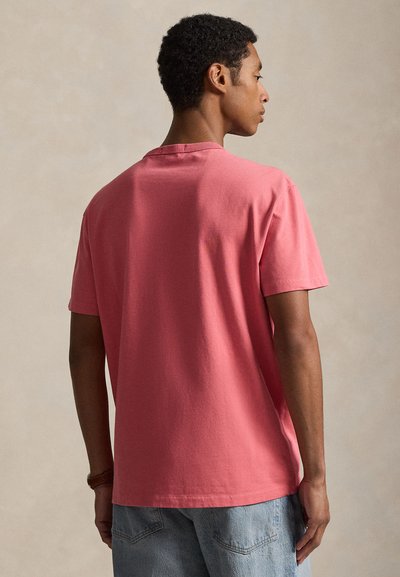 Άνδρας που στέκεται με την πλάτη γυρισμένη, φοράει απλό ροζ κοντομάνικο μπλουζάκι και ανοιχτό μπλε τζιν παντελόνι, μπροστά σε ουδέτερο φόντο.