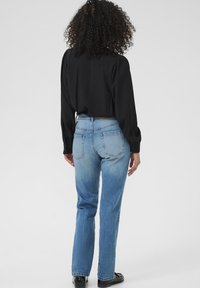 Vrouw met krullend haar, gekleed in een zwarte blouse met lange mouwen, lichtblauwe hoge taille jeans en zwarte schoenen, staand met haar rug naar de camera gericht.