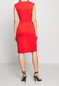 Červené, přiléhavé šaty s krátkými rukávy, délka ke kolenům a hladká textura. Nosené s černými vysokými sandály na podpatku, viditelným detailem bočního rozparku.