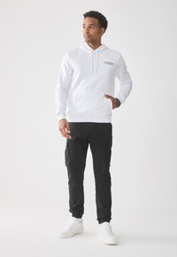 Uomo in felpa bianca con cappuccio, pantaloni cargo neri, sneakers bianche, che guarda di lato con una mano in tasca e l'altra rilassata.