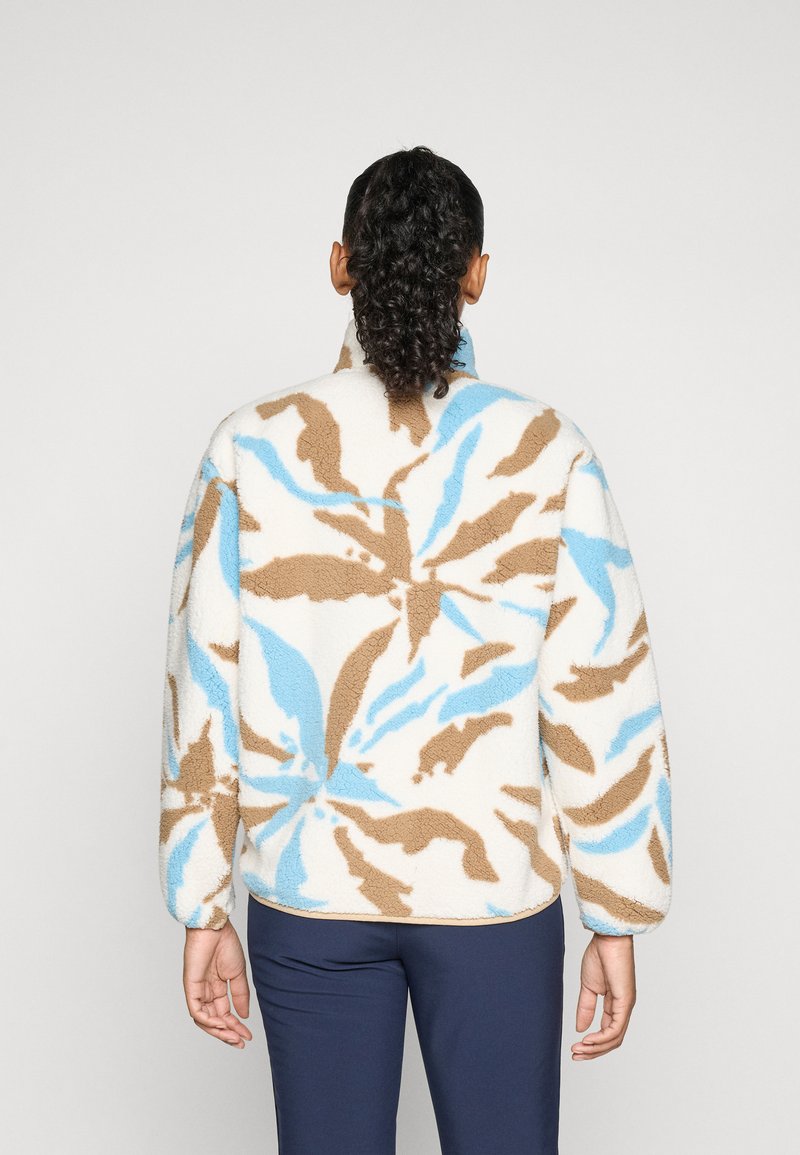 Osoba z kręconymi włosami związanymi w kucyk, ubrana w biały polarowy sweter z niebieskimi i beżowymi abstrakcyjnymi wzorami kwiatowymi, stojąca plecami na jednolitym tle.