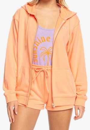 Žena oblečená v broskvově zbarvené mikině na zip a šortkách, přes fialový top s oranžovým nápisem „sunshine“ a grafikou palmy.