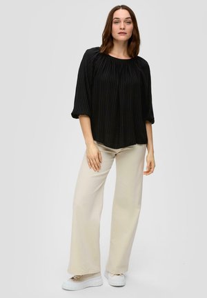 Blouse noire texturée à manches trois-quarts et col rond, associée à un pantalon large beige clair et des baskets à plateforme blanches.