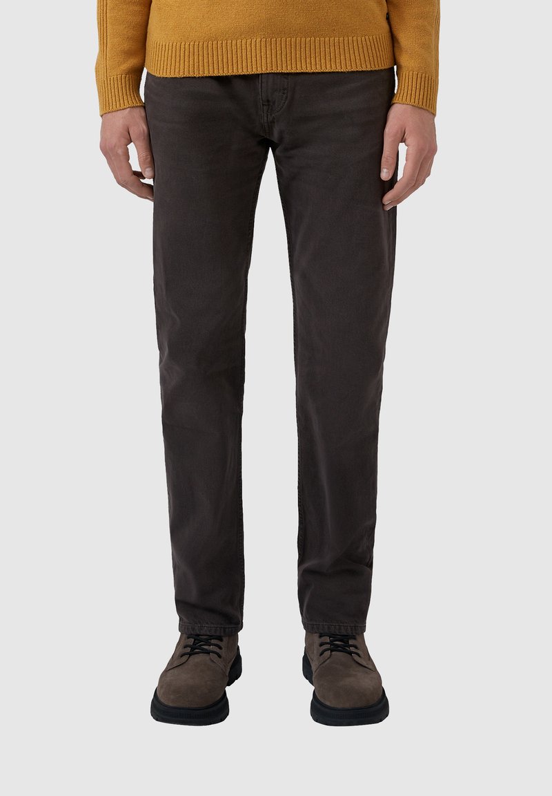 Osoba v tmavohnedých nohaviciach, hnedých semišových šnurovacích topánkach a horčicovožltom svetri stojí na svetlosivej pozadí.