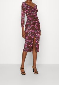 Vestido floral marrón de manga larga, con diseño fruncido y abertura lateral. Presenta patrones florales en rosa y azul sobre una tela ligera, combinado con tacones negros.