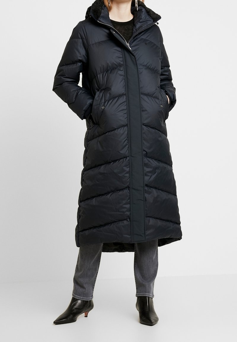 Long navy puffer coat with a zip closure, hood, and side pockets. Features a quilted texture and a straight cut design.