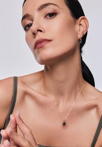 Złoty naszyjnik z owalnym czarnym zawieszką, noszony z szarym topem na ramiączkach. Modelka ma elegancką fryzurę i minimalny makijaż. Skoncentruj się na detalach biżuterii.