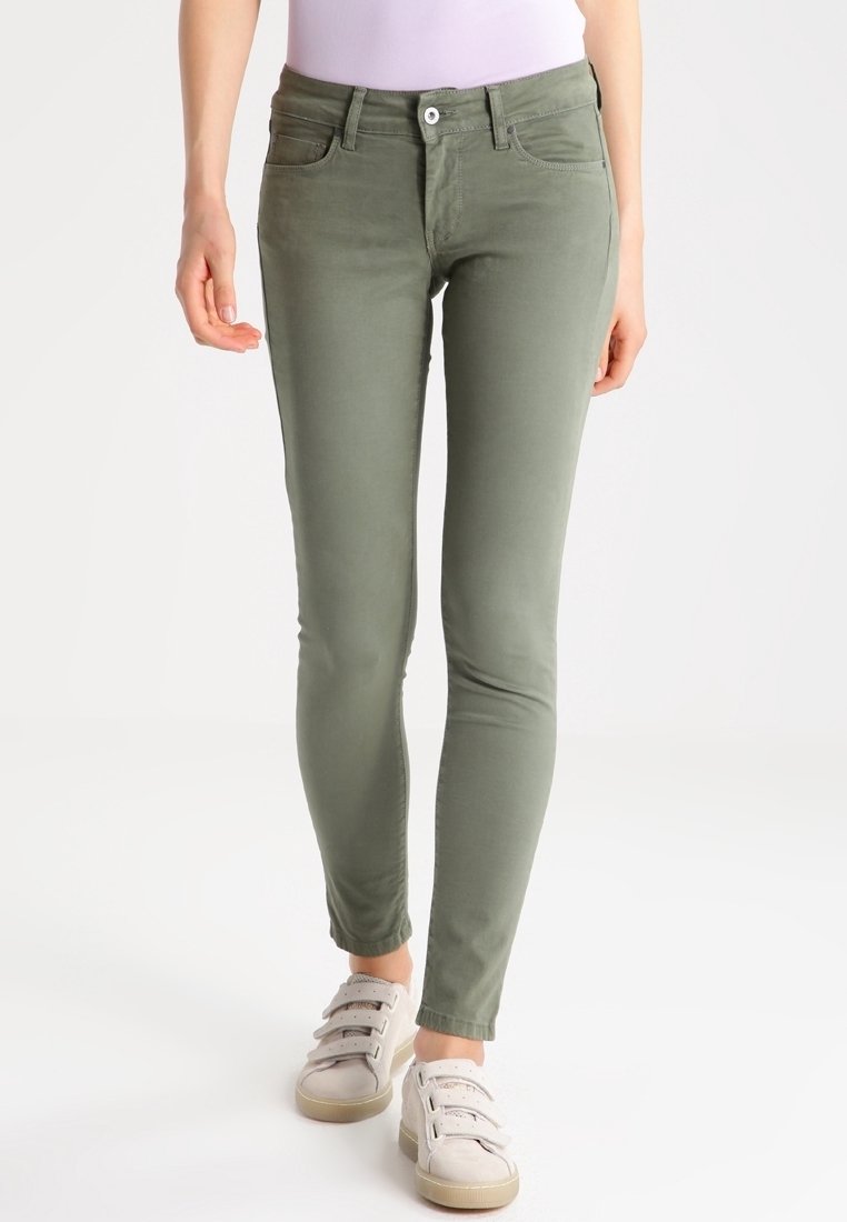 Jeans skinny verde oliva en algodón elástico con cinco bolsillos, trabillas para cinturón, cierre frontal con botón y cremallera, combinados con zapatillas informales.