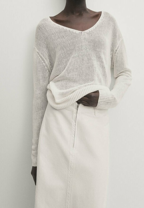 Personne à la peau foncée portant un pull blanc en maille ample, rentré dans une jupe blanche taille haute avec une fermeture éclair à l'avant.