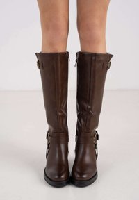 Bottes hautes en cuir marron à finition lisse, avec fermetures éclair latérales et boucles décoratives. Semelles en caoutchouc noir pour la stabilité.