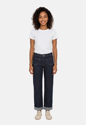 Donna con capelli ricci che indossa una t-shirt bianca, jeans scuri risvoltati e sneakers beige, in piedi davanti a uno sfondo chiaro e semplice.