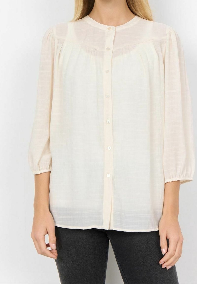 Blouse à manches longues couleur crème clair avec fermeture boutonnée, col rond et manches froncées. Texture transparente avec des rayures subtiles.