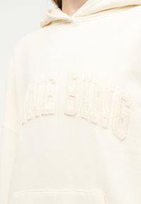 Κοντινό πλάνο κρεμ-χρωματισμένου φούτερ με κουκούλα με ανάγλυφα γράμματα "MAKE OWNG" στο στήθος και ορατή τσέπη στο μπροστινό μέρος στο κάτω μέρος.