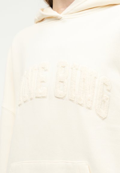 Gros plan sur un sweat à capuche couleur crème avec les lettres en relief "MAKE OWNG" sur la poitrine et une poche avant visible en bas.