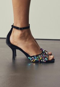 Sandali con tacco alto in velluto nero, caratterizzati da una parte superiore in paillettes con paillettes rotonde multicolori, design open toe e cinturino alla caviglia regolabile.