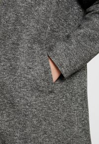 Cappotto grigio testurizzato con un dettaglio della tasca visibile; il tessuto appare morbido con un sottile motivo melange. La manica è leggermente arrotolata.