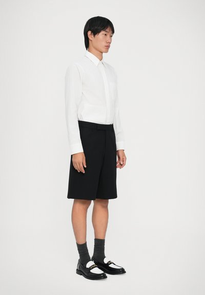 Mand iført hvid langærmet skjorte, sorte knælange shorts, mørkegrå sokker og sort-hvide loafers, der står mod en ensfarvet baggrund.