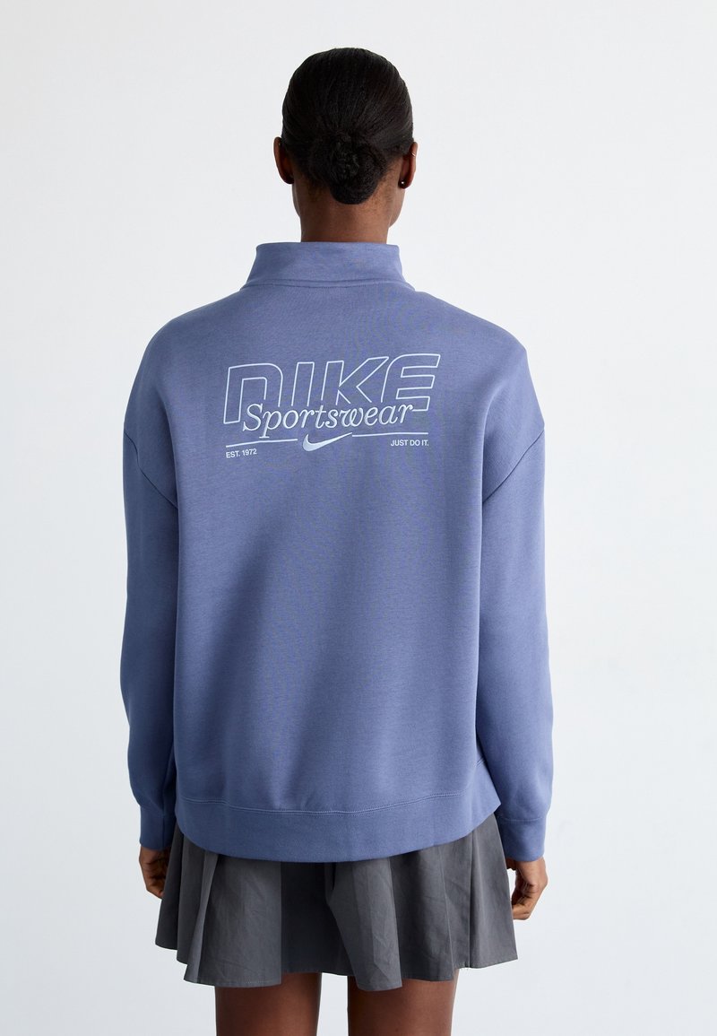 Ljusblå Nike-tröja med hög krage, med vit logotext på baksidan. Mjuk textur och avslappnad passform. Kombineras med en grå kjol.