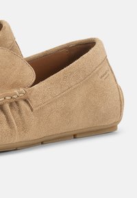 Μπεζ μοκασίνι από σουέτ με ραφές moc toe, σχέδιο που φοριέται εύκολα χωρίς κορδόνια και επίπεδη λαστιχένια σόλα, φωτογραφημένο σε λευκό φόντο.