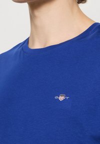 Personne portant un t-shirt bleu à col rond avec un petit logo GANT représentant un bouclier et des étoiles sur la poitrine gauche.