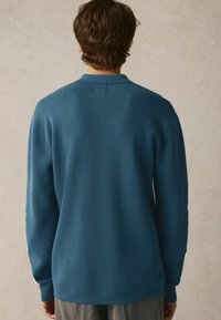 Mørkegrøn strikket sweater med krave, lange ærmer og ribbede manchetter. Glat tekstur med diskret sømdetalje ved halsudskæringen.
