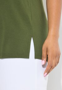 Olivově zelená bavlněná košile s bočním rozparkem, ukazující detaily šití. Ruka s růžovými nehty spočívá na bílé látce pod košilí.