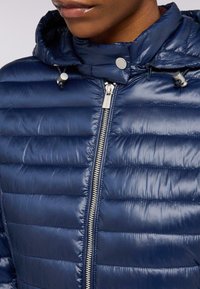 Giacca puffer blu navy con motivo a righe orizzontali, chiusura con zip metallica e cappuccio con cordino. Tessuto liscio e lucido.