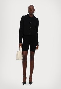Maglione nero corto con grande colletto, dettagli con bottoni, abbinato a pantaloni ciclisti neri. La modella tiene una piccola borsa crema e indossa tacchi neri.