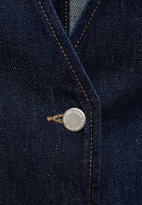 Ciemnoniebieska dżinsowa tkanina z widoczną teksturą i szwami, z metalowym przyciskiem w kolorze srebra z wygrawerowanym napisem oraz brązowym detalem z nici.