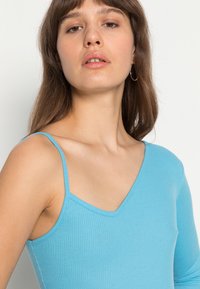 Top azul com pregas, decote em V e alças finas; apresenta um design ajustado com uma manga longa e uma textura suave.