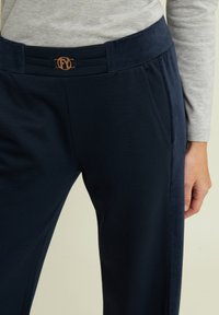 Pantaloni blu navy con una texture liscia, caratterizzati da una fibbia decorativa in vita e tasche laterali. Pannello laterale in un materiale contrastante.