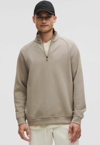 Beige pullover sweatshirt met een kwart-rits kraag, raglan mouwen en geribbelde manchetten, gedragen met een lichtgekleurde onderhemd en lichte broek.