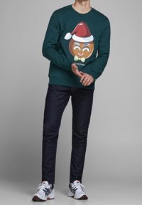 Sudadera verde con un gráfico de galleta de jengibre que lleva un sombrero de Santa, una cara sonriente y un pajarita, combinada con jeans oscuros y zapatillas.