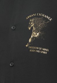 Μαύρο ύφασμα με χρυσό μοτίβο αλόγου και κείμενο "Armani Exchange" και "Ελευθερία νου, σώματος και πνεύματος" κοντά σε μαύρους κουμπιά.