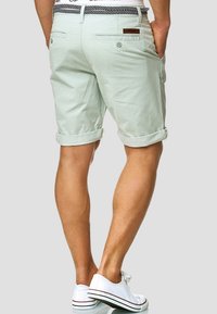 Pantalones cortos de algodón verde claro con mangas enrolladas, bolsillos traseros y un parche con el logo en la cinturilla, combinados con zapatillas blancas.