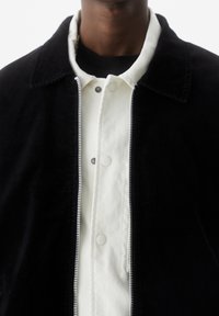 Giacca in corduroy nera con una camicia bianca con colletto sotto, dotata di chiusura con zip, bottoni a pressione e dettagli di cucitura visibili.