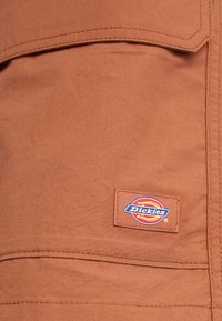 Tela marrón con una textura suave, con una etiqueta de parche que presenta el logo "Dickies". Bolsillo visible con detalle de costura contorneada.