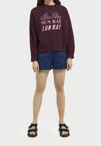 Sudadera burdeos con un gran texto "Rayos de Sol", combinada con pantalones cortos azul marino. El modelo lleva sandalias negras. El diseño presenta un corte holgado y cuello redondo.