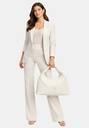 Donna che indossa un completo color crema e una canottiera, con una grande borsa bianca in mano, in piedi davanti a uno sfondo chiaro e semplice.