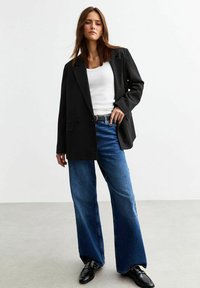 Blazer nero dalla vestibilità su misura, indossato sopra una canotta bianca. Abbinato a jeans in denim blu a gamba larga e scarpe nere, creando un look casual.