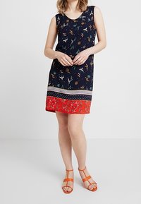 Ärmlös marinblå klänning med blommönster, röd mönstrad nederkant och kontrasterande randdetaljer. I kombination med orange sandaler med remmar.