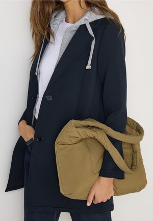 Femme portant un manteau bleu marine et un sweat à capuche gris tient un grand sac fourre-tout matelassé vert olive avec des poignées courtes.